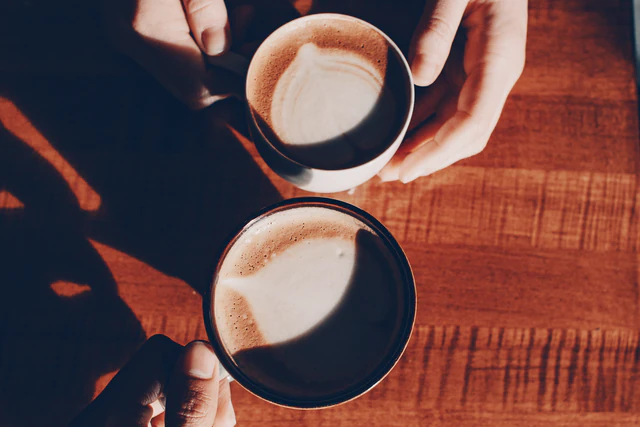 Manos sosteniendo dos tazas de café con leche sobre una mesa de madera al sol
